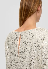 Top en sequins avec un col rond, présentant un détail en forme de clé à l'arrière. Principalement argenté avec des touches de sequins bleus.