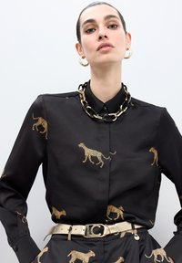 Mujer viste una camisa y pantalones negros con estampado de leopardo dorado, un collar grueso de cadena en negro y dorado, pendientes de aro dorados y un cinturón metálico dorado.