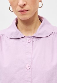 Ανοιχτό μωβ πουκάμισο με γιακά και κλείσιμο με κουμπιά στο μπροστινό μέρος, υφασμάτινη υφή και κοντά, μαζεμένα μανίκια. Διαθέτει κουμπιά σε τόνους ροζ που ταιριάζουν.