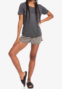 La camisa gris de manga corta presenta un diseño de texto, combinada con pantalones cortos gris claro. El modelo lleva sandalias negras de tipo deslizante, mostrando un tatuaje en la pierna en el tobillo derecho.