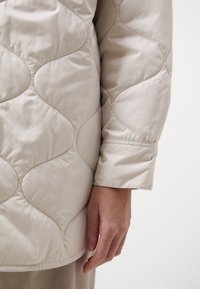 Jachetă bej căptușită cu un design matlasat, prezentând modele rotunjite, asemănătoare cu valuri. Textură netedă cu mâneci scurte, croite.