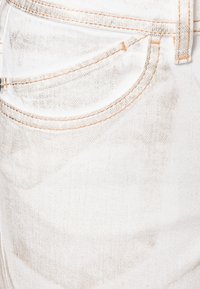 Vit denimtyg med ett urblekt mönster, med orange sömmar längs fickorna och midjebandet. Slät textur, närbild.