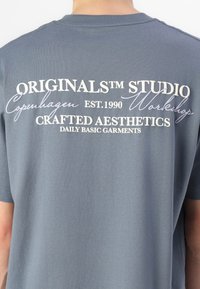 Grå bomulls t-shirt med vit text på baksidan: "ORIGINALS STUDIO Köpenhamn EST.1990" och "CRAFTED AESTHETICS" med kursiv accent.