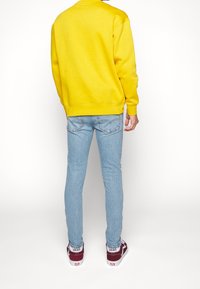 Gul sweatshirt med en avslappnad passform, ribbade ärmslut, matchad med ljusblå skinny jeans. Skorna är maroon Vans med vita sulor.