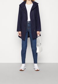 Cappotto impermeabile blu navy con cappuccio, maniche lunghe e zip frontale. Indossato sopra una camicia bianca e abbinato a jeans blu e sneakers bianche.