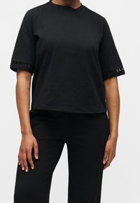 Personne portant un haut noir à manches courtes avec bordure en dentelle sur les manches et un pantalon noir, se tenant devant un fond clair uni.
