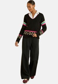 Melnā adītā džemperis ar V-veida kaklu, kuram uz piedurknēm ir rozā un tirkīza svītras, un zebraina apmale; apvienots ar melnām platajām biksēm.