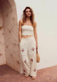 Top corto beige senza spalline abbinato a pantaloni larghi color crema con ricami floreali e dettagli a intaglio, accessoriato con una borsa intrecciata.