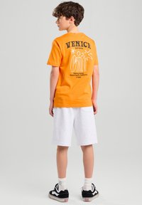 T-shirt in cotone arancione con stampa nera che presenta "VENICE surf shop" e un design grafico. Abbinato a pantaloni corti bianchi e scarpe da ginnastica nere.