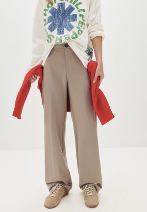 Persona que lleva pantalones beige de pierna ancha, zapatillas beige, una sudadera blanca con estampado azul y verde, sosteniendo un suéter rojo por los lados.