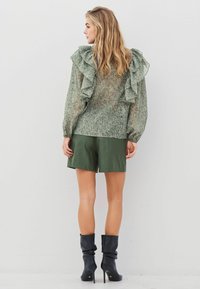 Grön volangblus med genomskinlig textur, parat med mörkgröna shorts och svarta avslappnade ankelstövlar, sedd bakifrån.