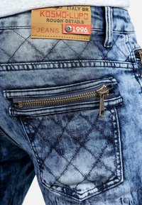 Close-up of blue acid-wash jeans pocket with zipper and brown leather label reading Kosmo Lupo 1996 Rough Details Italy BR.