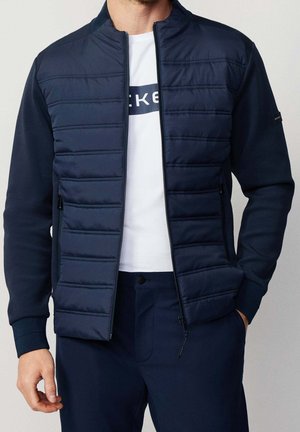 Homme portant une veste matelassée bleu marine sur une chemise blanche et un pantalon bleu marine assorti, debout avec une main dans la poche.
