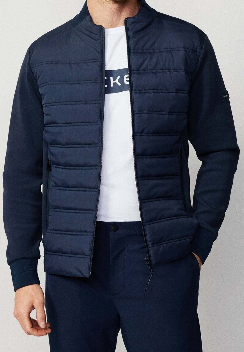 Homme portant une veste matelassée bleu marine sur une chemise blanche et un pantalon bleu marine assorti, debout avec une main dans la poche.