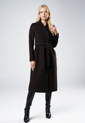 Blondynka ubrana w długi czarny płaszcz z paskiem i czarne kozaki do kolan, stojąca na tle jednolitego jasnoszarego tła.