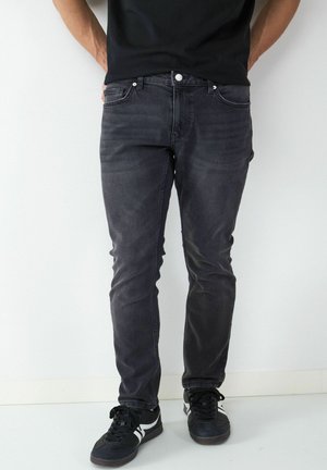 Mann trägt verwaschene schwarze Slim-Fit-Jeans und schwarze Sneaker mit weißen Streifen, steht vor einer schlichten weißen Wand.