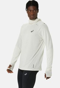 Langsleeve sporttop in lichtbeige, met een coltrui, ventilatiegaten op de schouders en duimgaten in de mouwen; gelabeld logo op de borst.