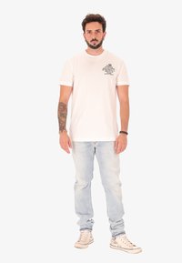 Uomo con un tatuaggio sul braccio sinistro che indossa una t-shirt bianca con grafica, jeans azzurri chiari, sneakers e braccialetti, in piedi contro uno sfondo bianco.