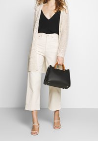 Schwarze Lederhandtasche mit strukturiertem Design, ausgestattet mit Bambusgriffen und goldfarbenen Metallakzenten. Das Modell trägt eine leichte Strickjacke und weite Hosen.