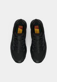 Zapatillas deportivas negras con una suela exterior de goma texturizada, parte superior de malla y cordones trenzados. Presentan una plantilla verde y amarilla con logotipo.