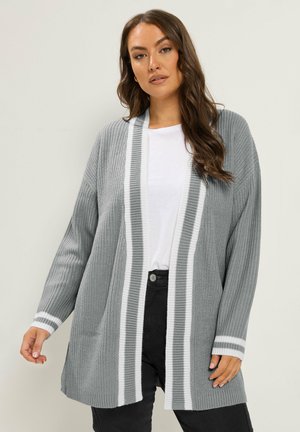 Femme portant un long cardigan gris tricoté avec des rayures blanches sur une chemise blanche et un pantalon noir devant un fond clair uni.