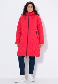 Rød lang puffjakke med et chevron-quiltet m�ønster, hætte, lynlås foran, sidelommer og sorte lynlåsdetaljer. Båret over blå jeans og hvide sneakers.