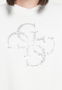 Valge dressipluus, mille keskosas on tekstuuriline, kristallidega kaunistatud logo, ribidega krae ja pehme kangas.