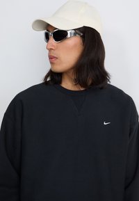 Oseba z dolgimi temnimi lasmi nosi črno majico Nike, belo baseball kapo in sončna očala s refleksivnim premazom, medtem ko pozira pred nevtralno ozadjem.