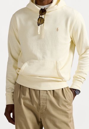 Mann trägt cremefarbene Kapuzenjacke mit Vorderfach und beigefarbene Hose, Sonnenbrille hängt an den Kapuzenbändern, und Armbanduhr am linken Handgelenk.