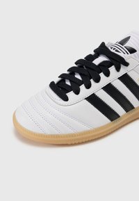 White athletic shoe with black stripes, textured upper, black laces, and a gum-colored sole featuring a patterned tread.