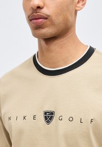 Κοντινό πλάνο ενός ατόμου που φοράει πουκάμισο γκολφ της Nike σε μπεζ απόχρωση με ασπρόμαυρο γιακά και λογότυπο με θέμα το γκολφ στο στήθος.