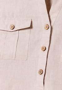 Partie de chemise en lin beige clair avec des boutons en bois à quatre trous et une poche à rabat sur le côté gauche.