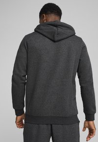 Man die met zijn rug naar de kijker staat, gekleed in een donkergrijze hoodie en bijpassende sweats tegen een effen lichtgrijze achtergrond.