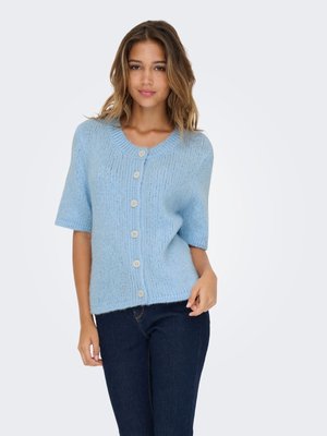 Ung kvinde iført lys blå kortærmet strikket cardigan med knapper, kombineret med mørkeblå jeans, stående mod en ensfarvet baggrund.