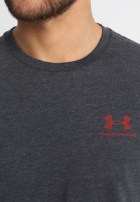 Šedé bavlněné tričko s žebrovaným límcem a krátkými rukávy, na levé části hrudi má červené logo Under Armour.