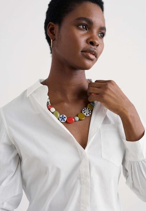 Multicolored bead necklace featuring round and oval stones in red, green, blue, and white, worn over a white collared shirt.