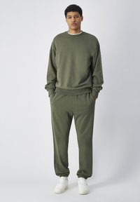 Felpa verde oliva con scollatura rotonda e maniche lunghe, abbinata a pantaloni jogger coordinati con polsini elasticizzati; sneakers bianche.
