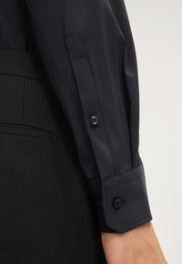 Camicia nera con una texture liscia e lucida, caratterizzata da una vestibilità su misura. Il polsino abbottonato presenta due bottoni neri e una finitura delle cuciture pulita.