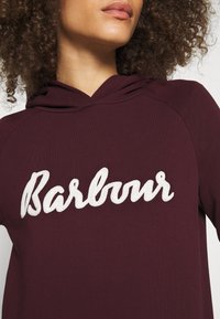 Bordeaux mikina s texturovaným bílým vyšívaným logem "Barbour". Obsahuje kapuci se stahovací šňůrkou a raglánové rukávy. Měkká a hladká tkanina.