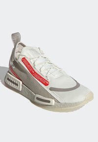 Lättviktig sneaker i vitt och beige med texturerade meshpaneler och grå accenter. Utrustad med röd branding och en gummisula för grepp.