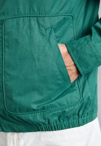 Grønn, lett jakke med synlig lomme, elastisk nederkant og jevn tekstur. Har enkel søm og klassisk design.