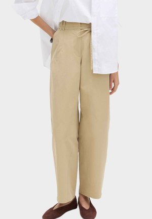 Beżowe spodnie z wysokim stanem i szerokimi nogawkami, z przednimi kieszeniami i paskiem do wiązania, w połączeniu z białą koszulą i brązowymi płaskimi butami.