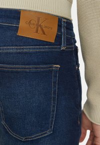 Vaqueros de mezclilla en azul oscuro con costuras claras, con una etiqueta de cuero marrón en la parte trasera de la cintura que dice "Calvin Klein Jeans".