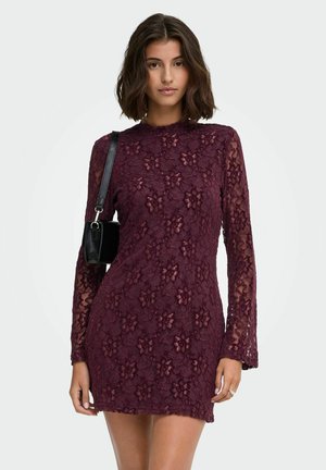 Femme aux cheveux bruns courts portant une robe en dentelle florale violet foncé à manches longues et portant un petit sac à bandoulière noir.