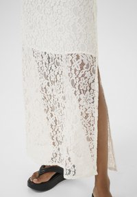 Jupe maxi en dentelle blanche avec un motif florale, dotée d'une fente latérale. Assortie avec des sandales plates noires, mettant en avant une semelle en plateforme texturée.