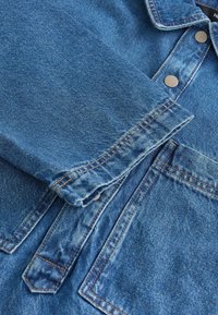 Denim jaka ar gaišu zilu nokrāsu, kurai ir noraksta apkakle, metāla pogas un redzamie šuvju detaļas uz piedurknēm un kabatām.