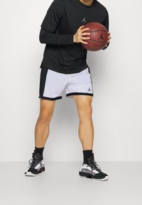 Svart långärmad tröja, vit och svart mesh basketshorts, och svarta och vita atletiska skor, hållande en basketboll.