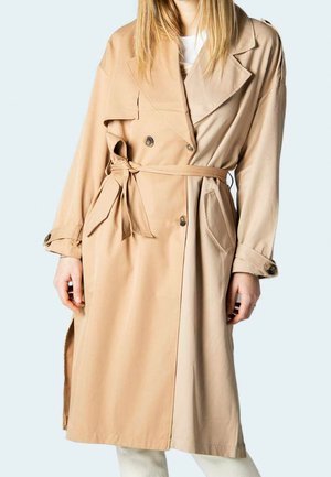 Femme portant un trench-coat beige longueur genou avec des boutons croisés, une ceinture à nouer à la taille et des poches latérales.