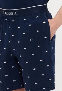 Sötétkék rövidnadrág, puha anyagból, rugalmas derékbőséggel, amelyen ismétlődő fehér Lacoste logó minta található.