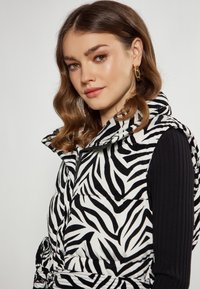 Zwart-wit gestreepte gewatteerde vest met hoge kraag en ritsluiting, gedragen over een zwart ribgebreid longsleeve shirt.